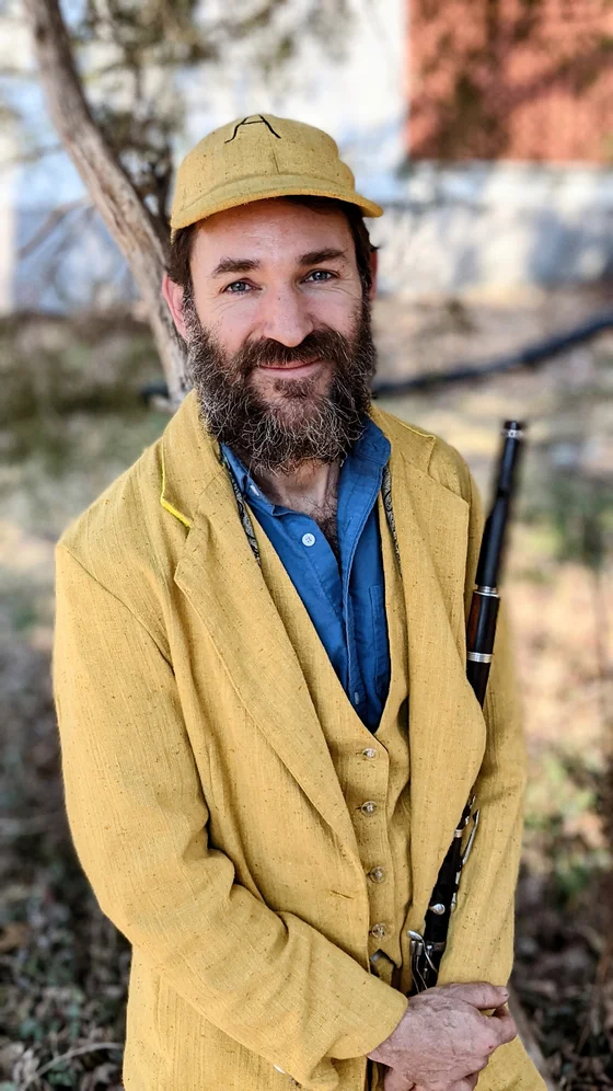 Aaron Olwell standing outdoors in a yellow work jacket, holding a wooden flute.